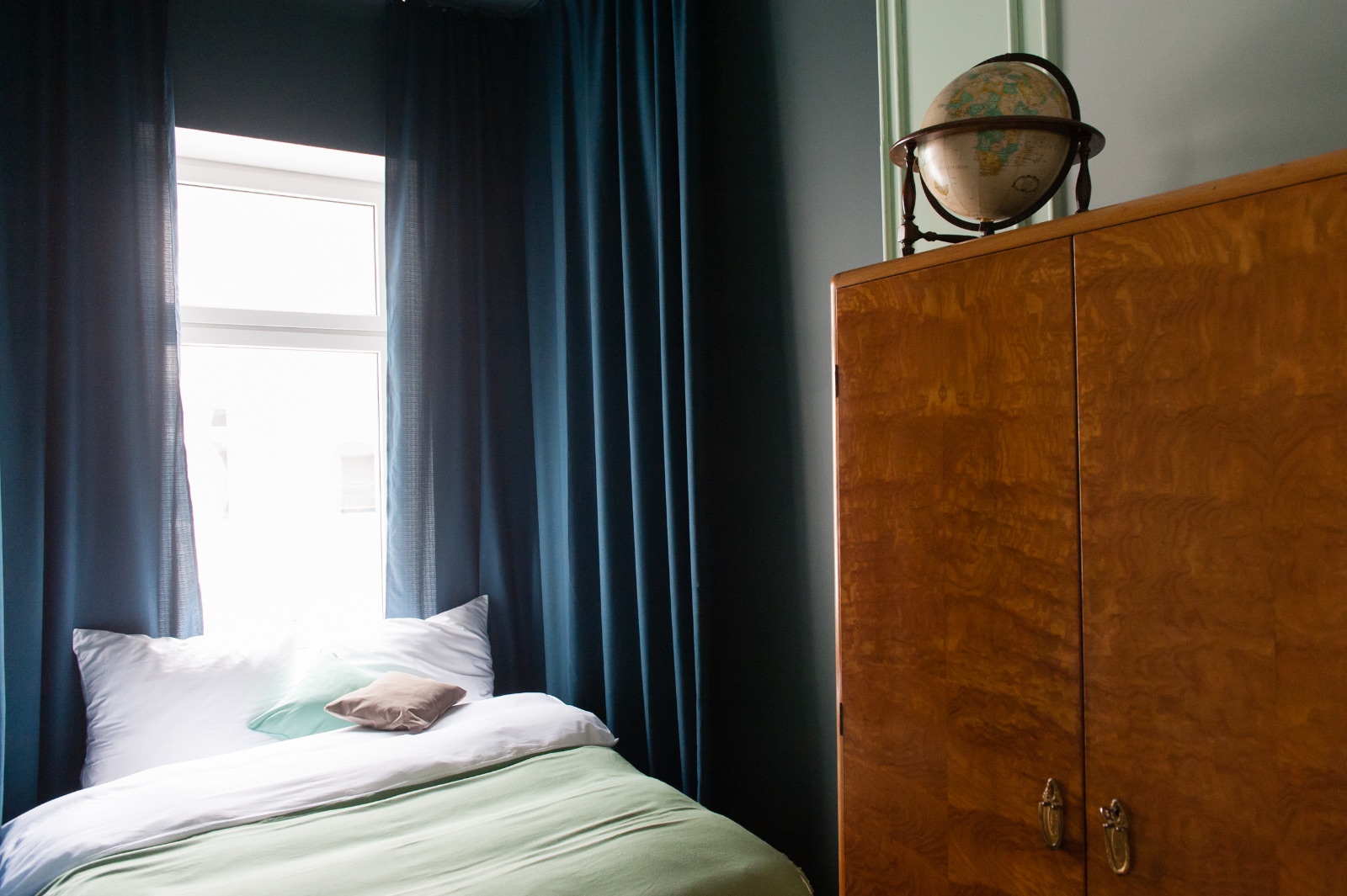 Furnished apartment in Vienna for a while with dark blue curtains, a bed with a mint green blanket and a burl wood cabinet with an old globe