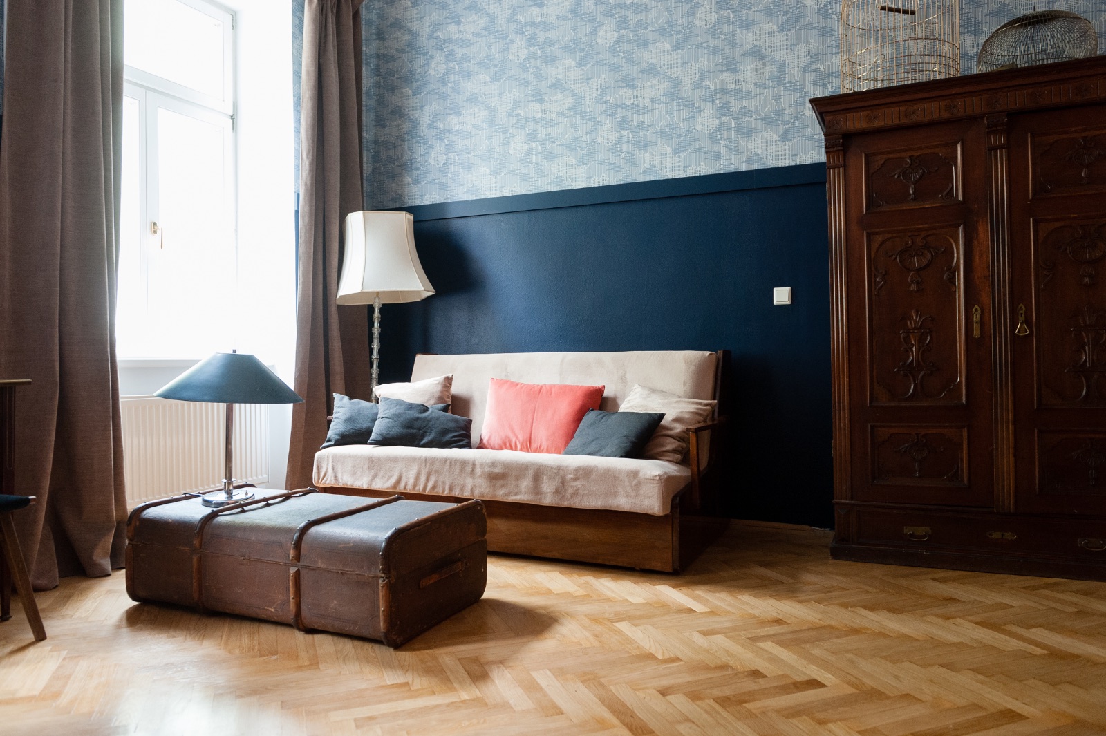 Living space of a furnished apartment in Vienna with an old leather trunk as a coffee table, a two tone blue wall, a carved wooden cabinet and herringbone parquet