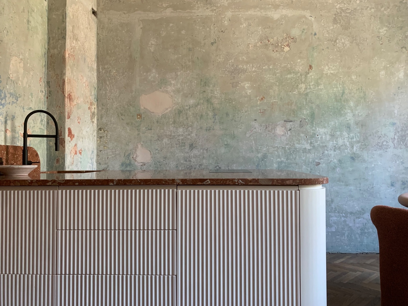 Geriffelte MDF-Kuecheninsel in gebrochenem Weiss mit rotgeaderter Marmorplatte und schwarzer Armatur vor einer rohen Kalkputzwand