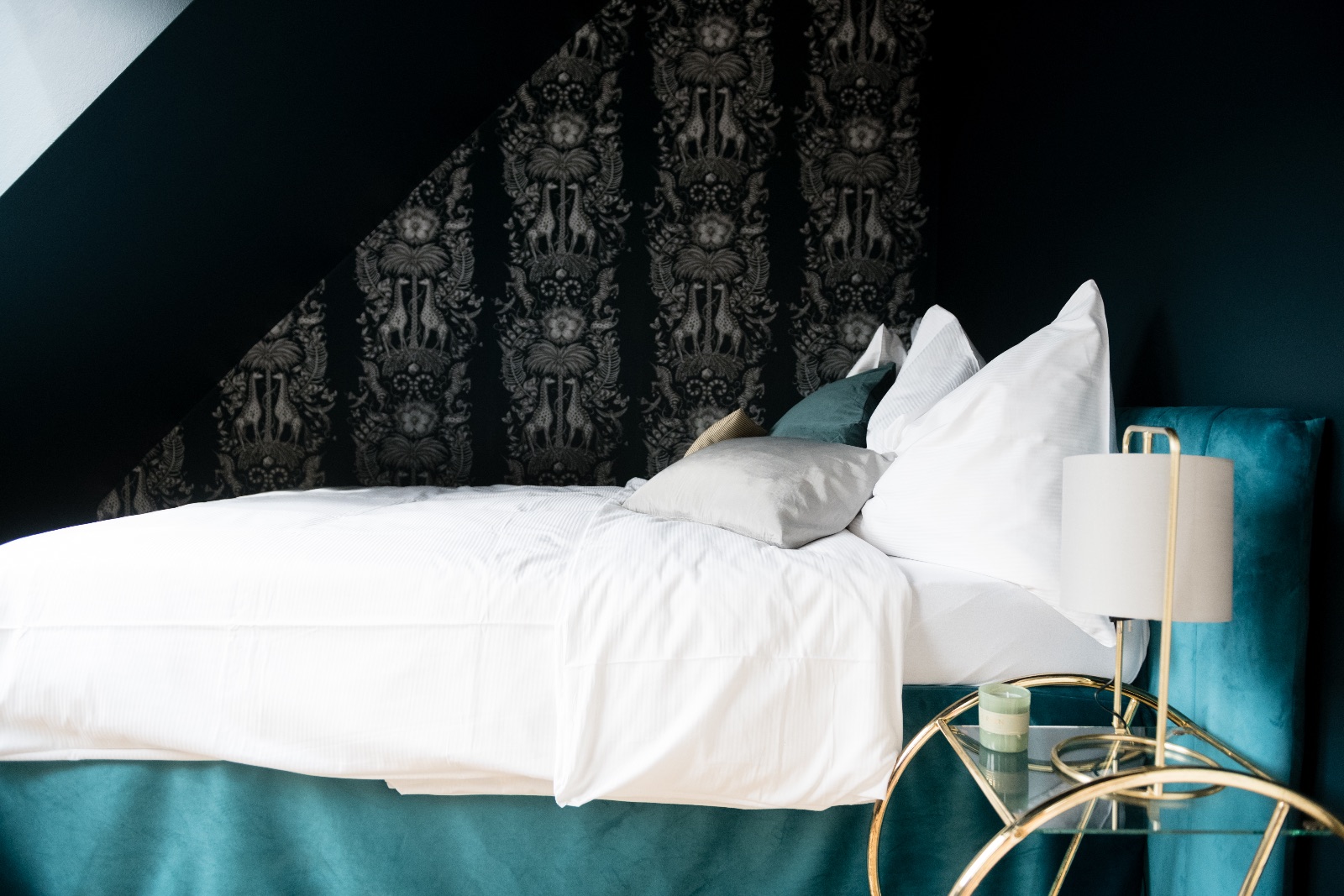 Chambre en bleu pétrole profond avec tête de lit en velours, linge de lit blanc, papier peint sombre à motif animalier et table d'appoint en laiton avec lampe