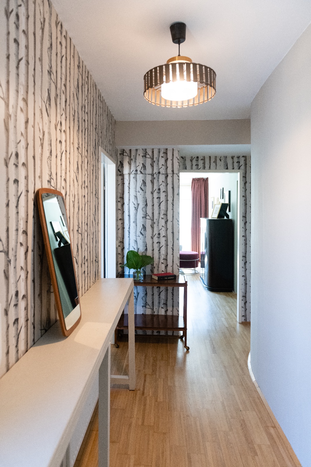 Möbliertes Apartment in Wien, Schlafzimmer mit Leinenbett und Pendelleuchte, Flur mit Birkentapete und Spiegel, Wohnraum mit grauem Samtsofa am Fenster