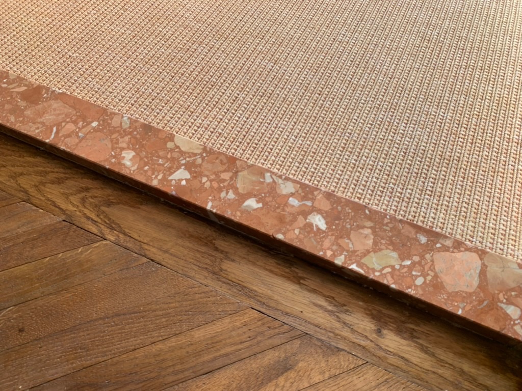 Transition de sol en marbre rouge entre parquet en chevron et tapis en sisal couleur sable a bordure brun rouge