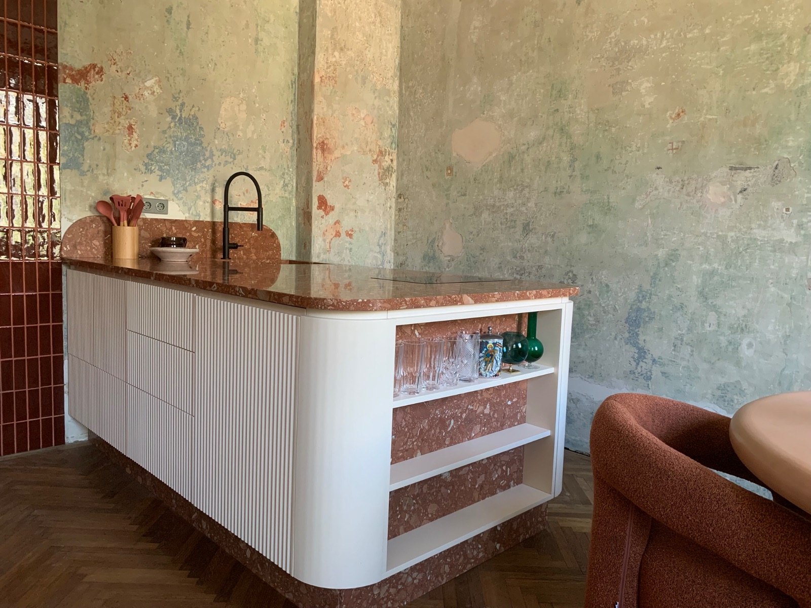 Ein modernes, möbliertes Apartment mit Terrazzo-Arbeitsplatte, weißem Regal mit braunen Einsätzen, Marmor-Details, gestaltet für Gastgeber auf Zeit. Im Hintergrund blättert die Wand ab in zarten Farben.